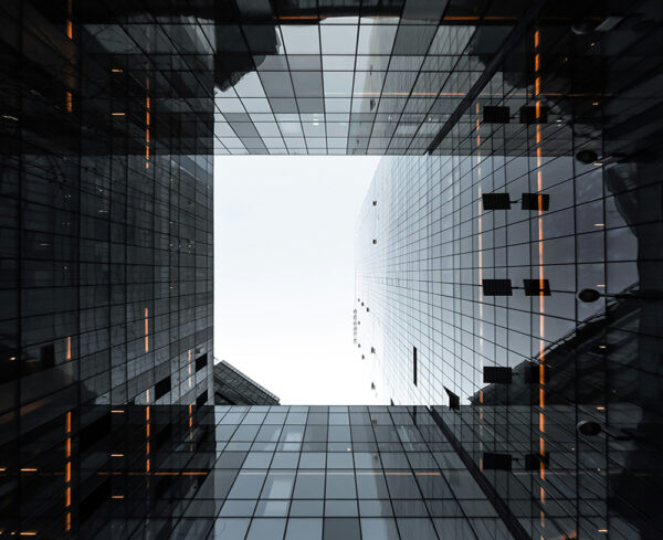 abstract, architectural perspective looking upward through a cluster of modern glass buildings. Sharp lines and reflective surfaces frame a bright opening of sky at the center