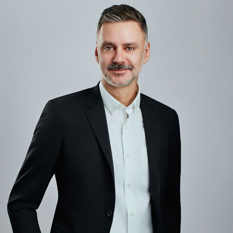 Chris Grimm looking straight into the camera with right hand on side elbow angled and left arm straight down at side.