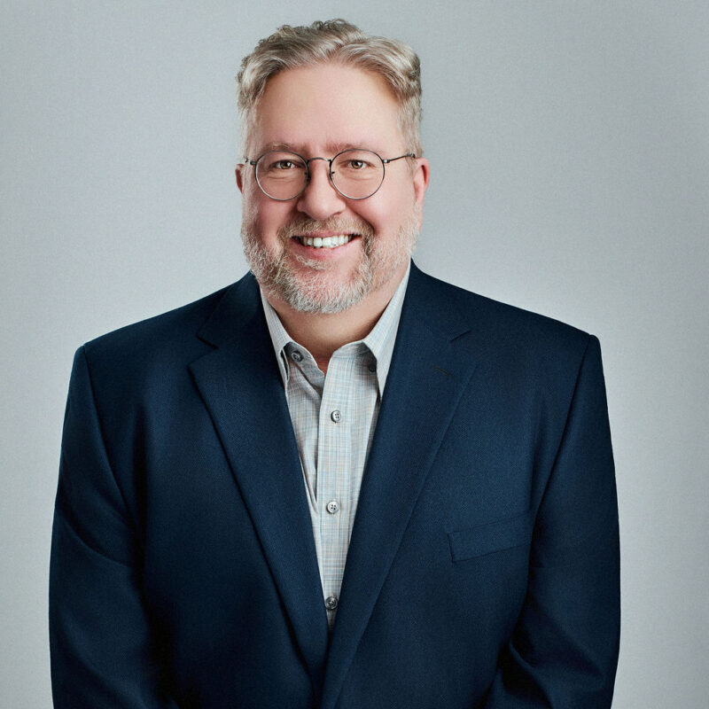 John Hicks looking straight into the camera smiling with arms at the sides.